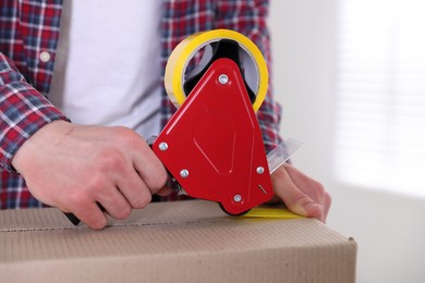 Photo of Man using dispenser with adhesive tape to seal cardboard box indoors, closeup