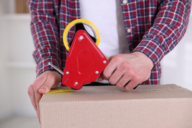 Photo of Man using dispenser with adhesive tape to seal cardboard box indoors, closeup