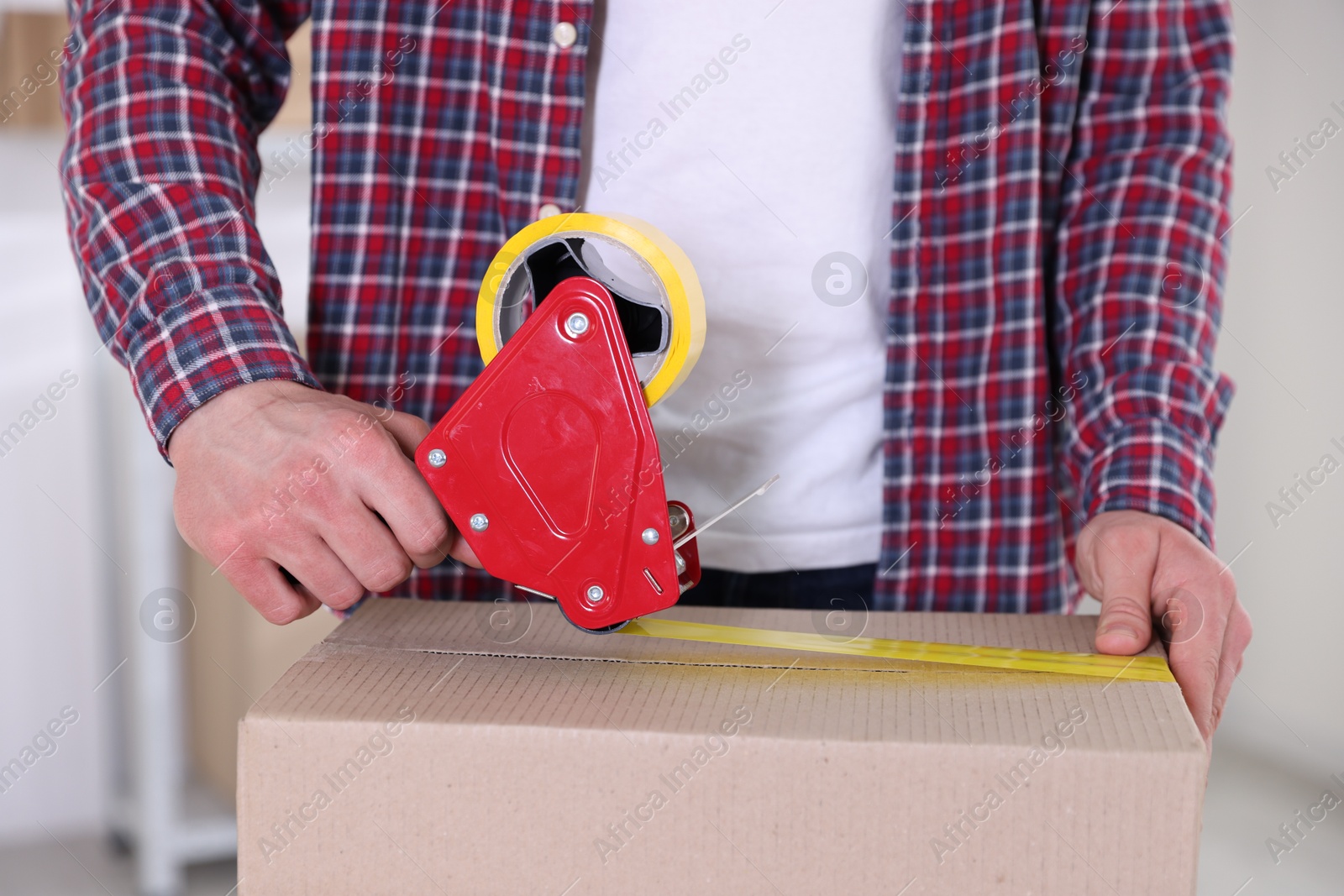 Man using dispenser with adhesive tape to seal cardboard box indoors, closeup Photo of Man using dispenser with adhesive tape to seal cardboard box indoors, closeup