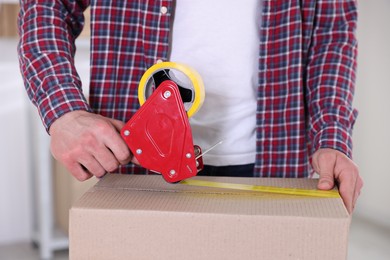 Photo of Man using dispenser with adhesive tape to seal cardboard box indoors, closeup