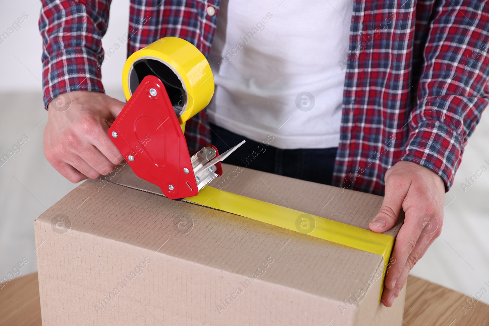 Man using dispenser with adhesive tape to seal cardboard box indoors, closeup Photo of Man using dispenser with adhesive tape to seal cardboard box indoors, closeup