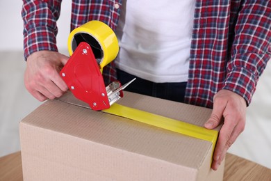 Photo of Man using dispenser with adhesive tape to seal cardboard box indoors, closeup