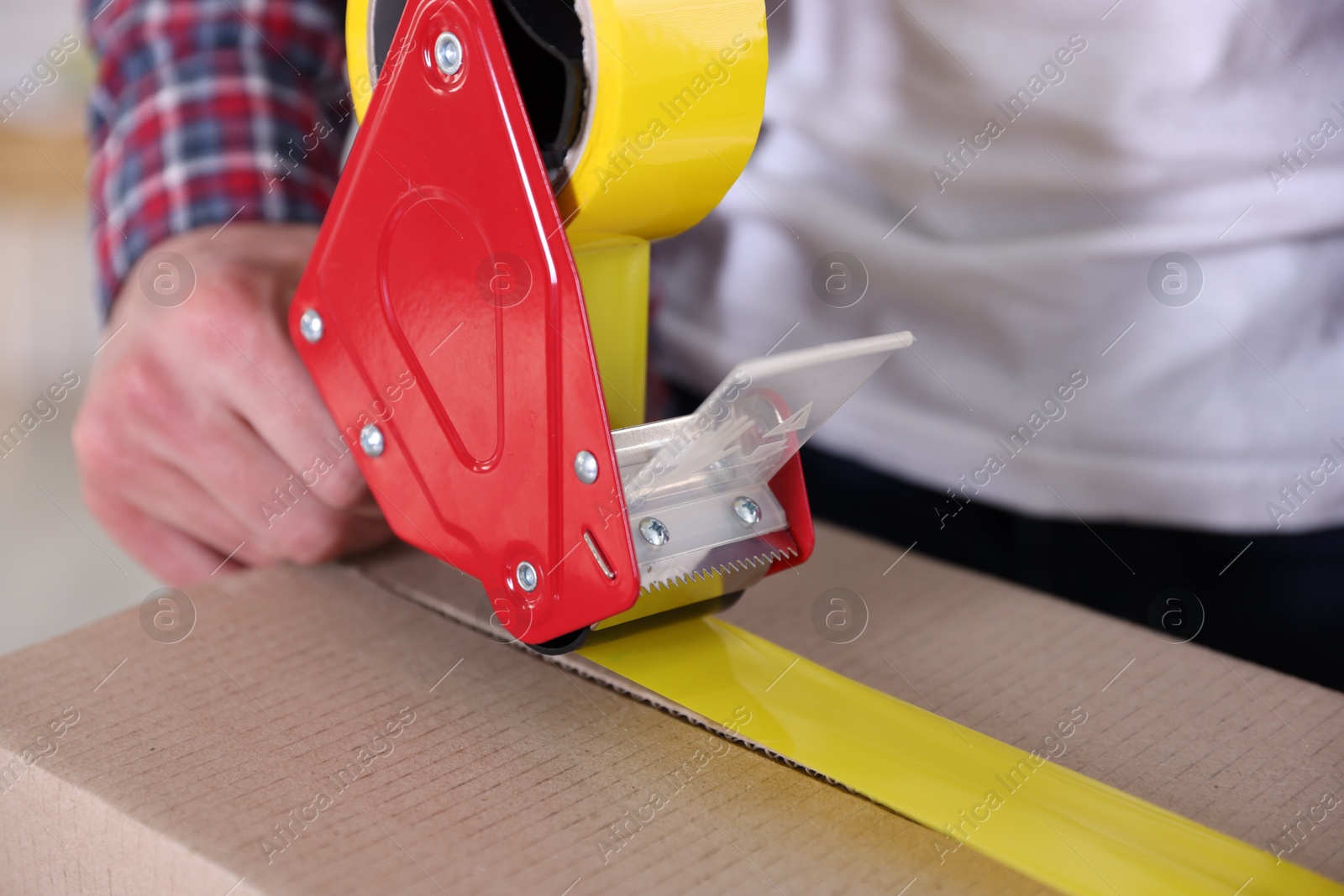 Man using dispenser with adhesive tape to seal cardboard box indoors, closeup Photo of Man using dispenser with adhesive tape to seal cardboard box indoors, closeup