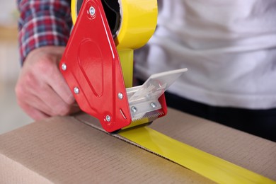 Photo of Man using dispenser with adhesive tape to seal cardboard box indoors, closeup