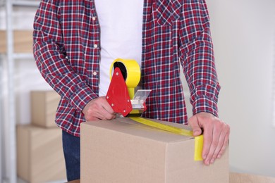 Photo of Man using dispenser with adhesive tape to seal cardboard box indoors, closeup