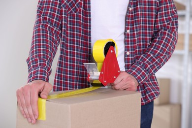Photo of Man using dispenser with adhesive tape to seal cardboard box indoors, closeup