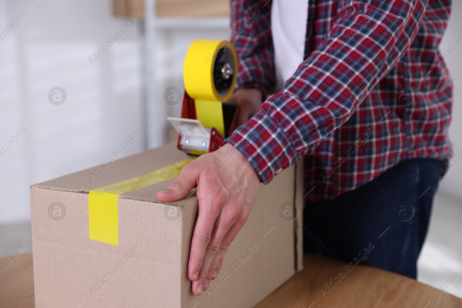 Man using dispenser with adhesive tape to seal cardboard box indoors, closeup Photo of Man using dispenser with adhesive tape to seal cardboard box indoors, closeup