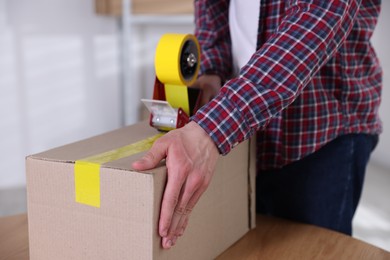Photo of Man using dispenser with adhesive tape to seal cardboard box indoors, closeup