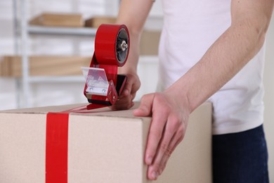 Photo of Man using dispenser with adhesive tape to seal cardboard box indoors, closeup