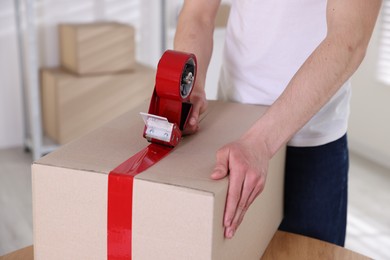 Photo of Man using dispenser with adhesive tape to seal cardboard box indoors, closeup