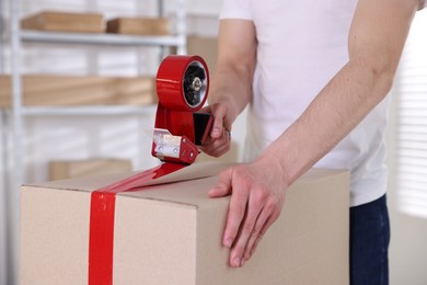 Photo of Man using dispenser with adhesive tape to seal cardboard box indoors, closeup