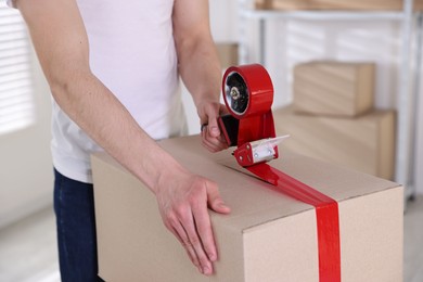 Photo of Man using dispenser with adhesive tape to seal cardboard box indoors, closeup