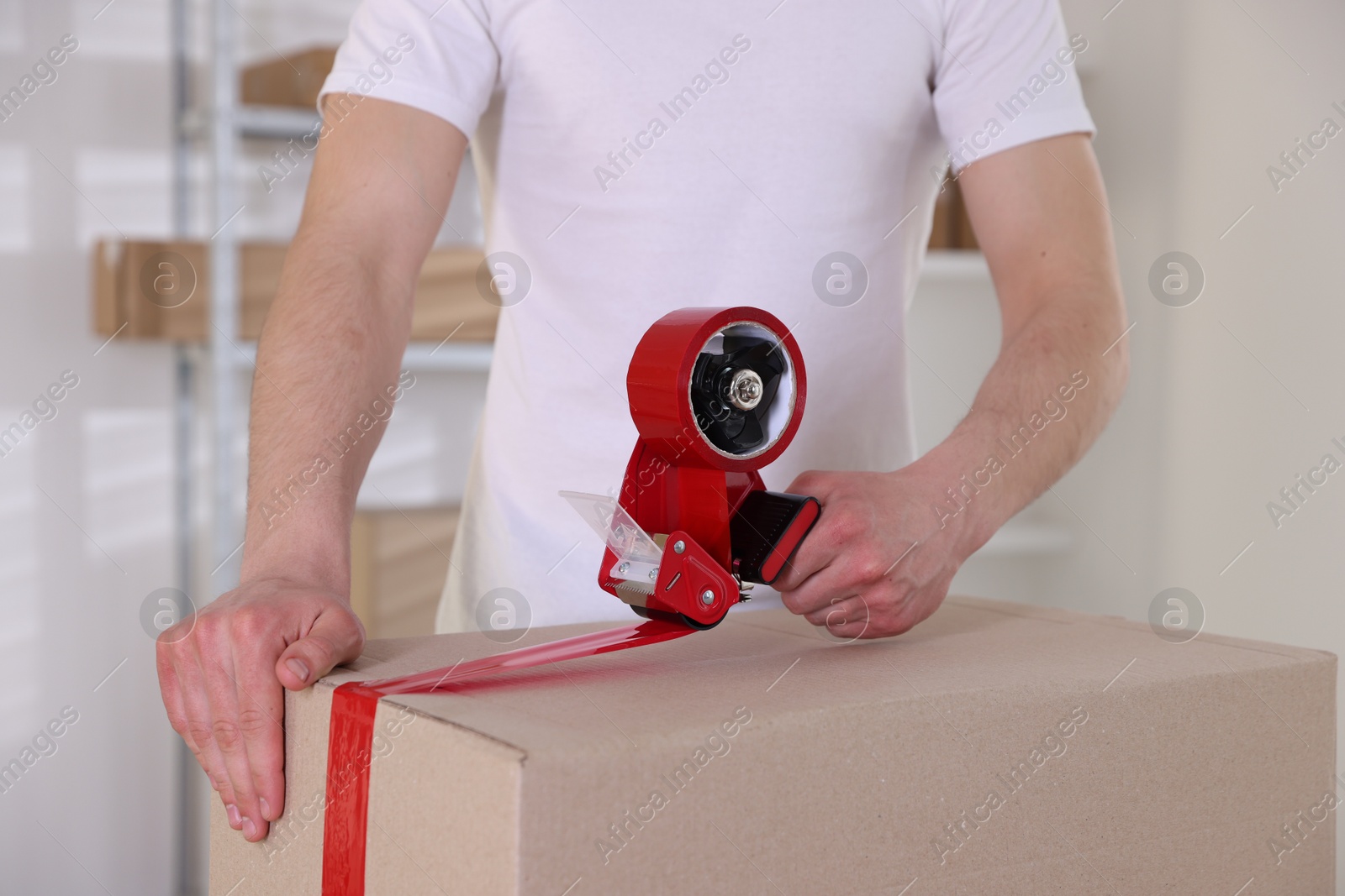 Man using dispenser with adhesive tape to seal cardboard box indoors, closeup Photo of Man using dispenser with adhesive tape to seal cardboard box indoors, closeup