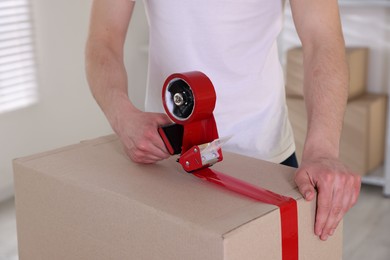 Photo of Man using dispenser with adhesive tape to seal cardboard box indoors, closeup
