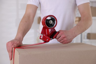 Photo of Man using dispenser with adhesive tape to seal cardboard box indoors, closeup