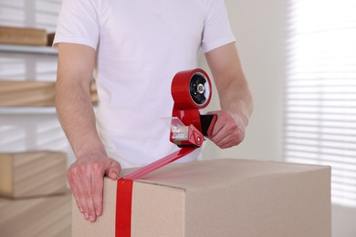 Photo of Man using dispenser with adhesive tape to seal cardboard box indoors, closeup