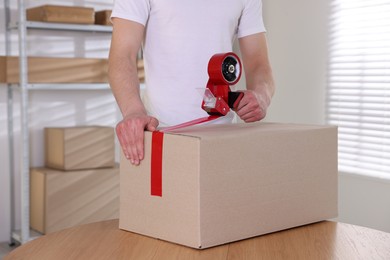 Photo of Man using dispenser with adhesive tape to seal cardboard box indoors, closeup