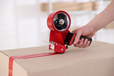 Photo of Man using dispenser with adhesive tape to seal cardboard box indoors, closeup