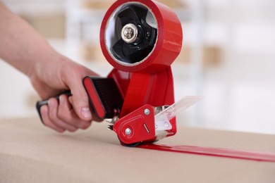 Photo of Man using dispenser with adhesive tape to seal cardboard box indoors, closeup