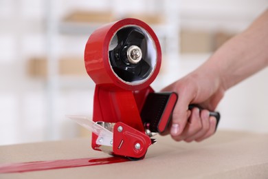 Photo of Man using dispenser with adhesive tape to seal cardboard box indoors, closeup