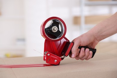 Photo of Man using dispenser with adhesive tape to seal cardboard box indoors, closeup