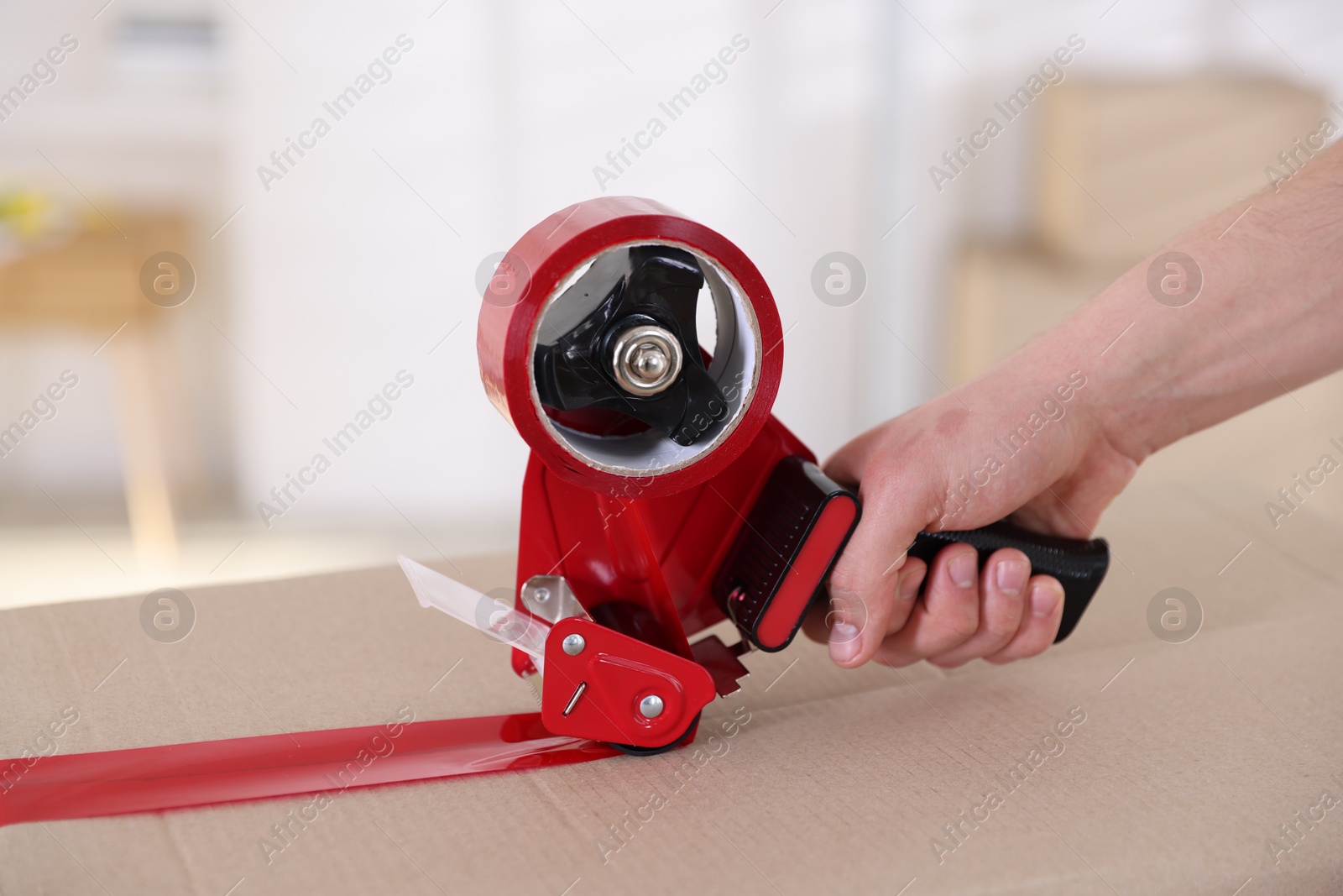 Photo of Man using dispenser with adhesive tape to seal cardboard box indoors, closeup