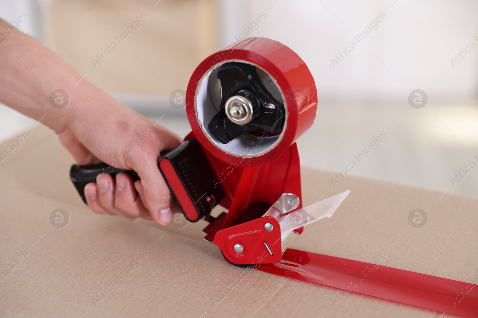 Photo of Man using dispenser with adhesive tape to seal cardboard box indoors, closeup