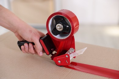 Photo of Man using dispenser with adhesive tape to seal cardboard box indoors, closeup