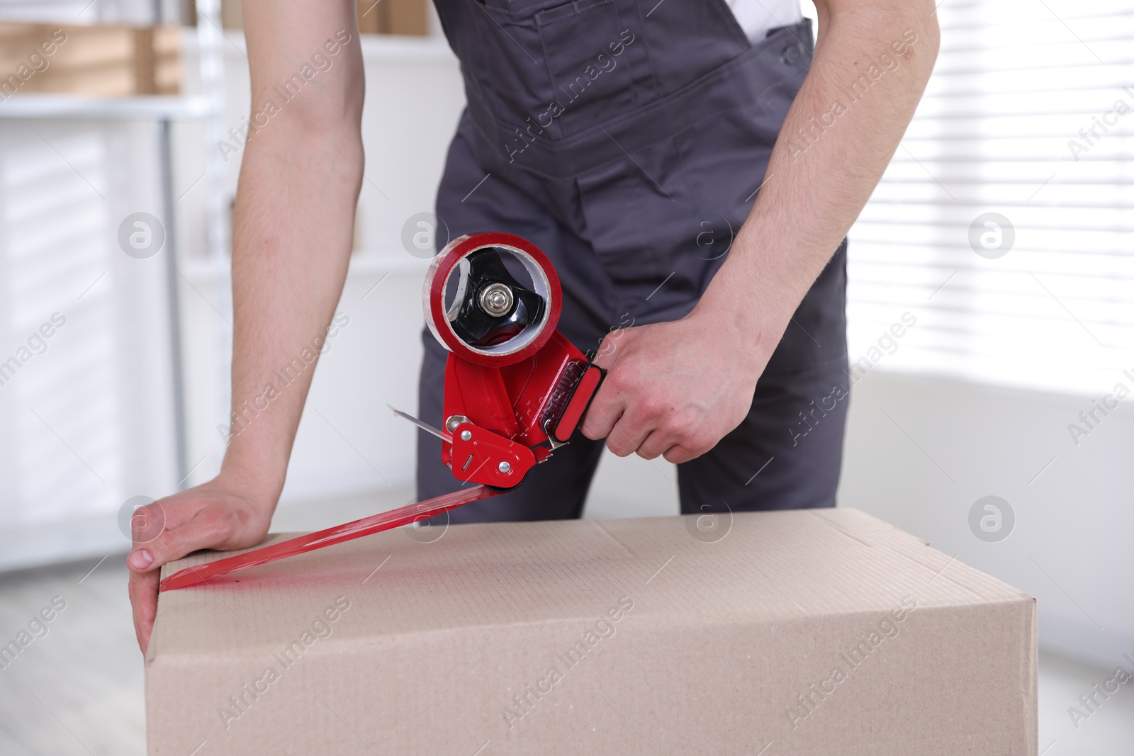 Photo of Man using dispenser with adhesive tape to seal cardboard box indoors, closeup