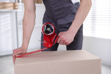 Photo of Man using dispenser with adhesive tape to seal cardboard box indoors, closeup