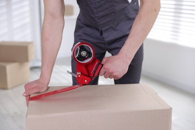 Photo of Man using dispenser with adhesive tape to seal cardboard box indoors, closeup