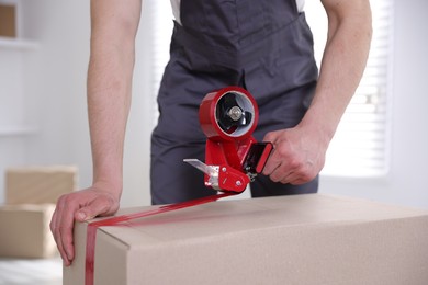 Photo of Man using dispenser with adhesive tape to seal cardboard box indoors, closeup