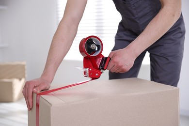 Photo of Man using dispenser with adhesive tape to seal cardboard box indoors, closeup