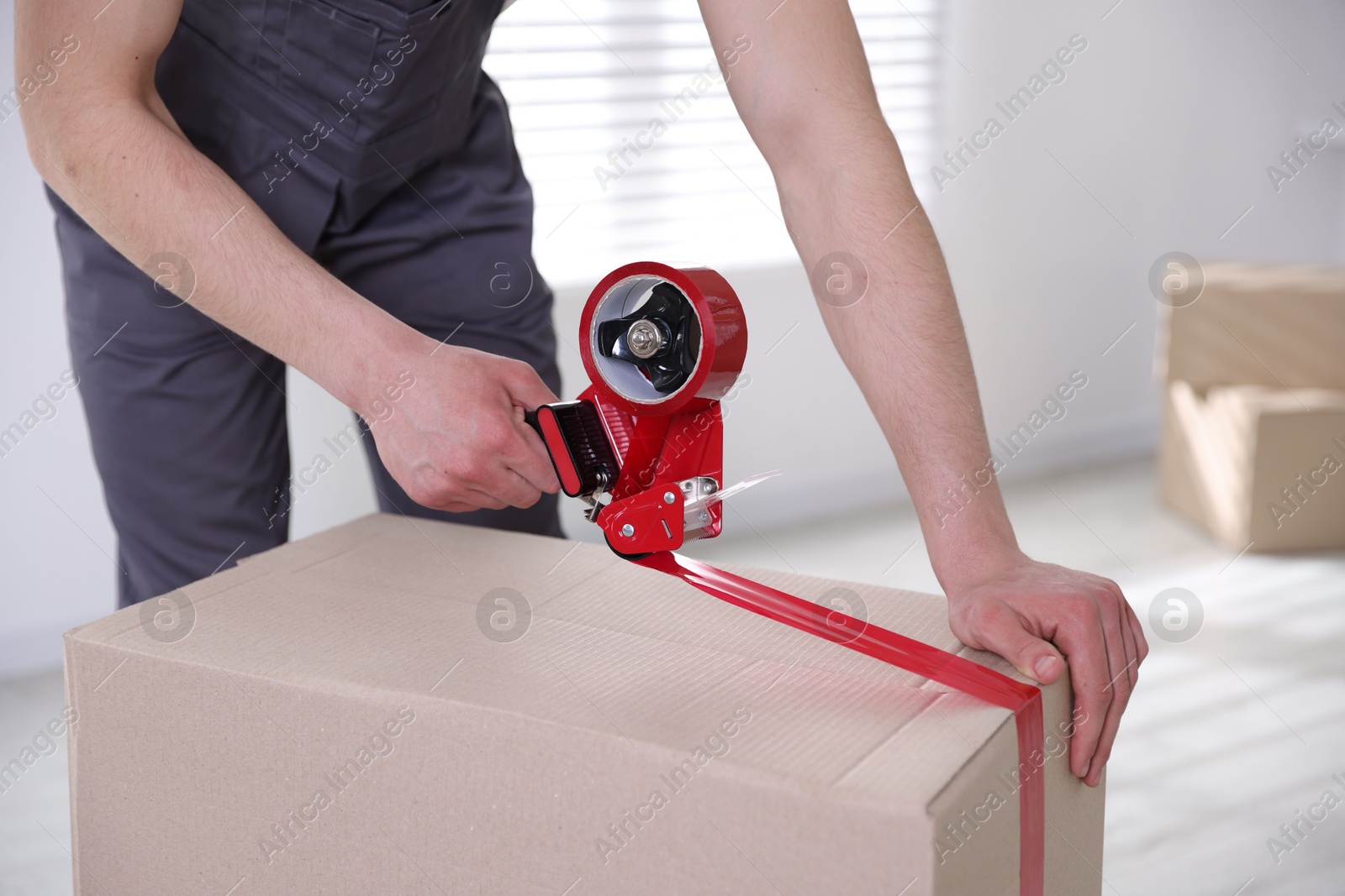 Photo of Man using dispenser with adhesive tape to seal cardboard box indoors, closeup