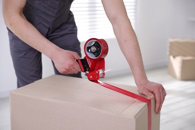 Photo of Man using dispenser with adhesive tape to seal cardboard box indoors, closeup