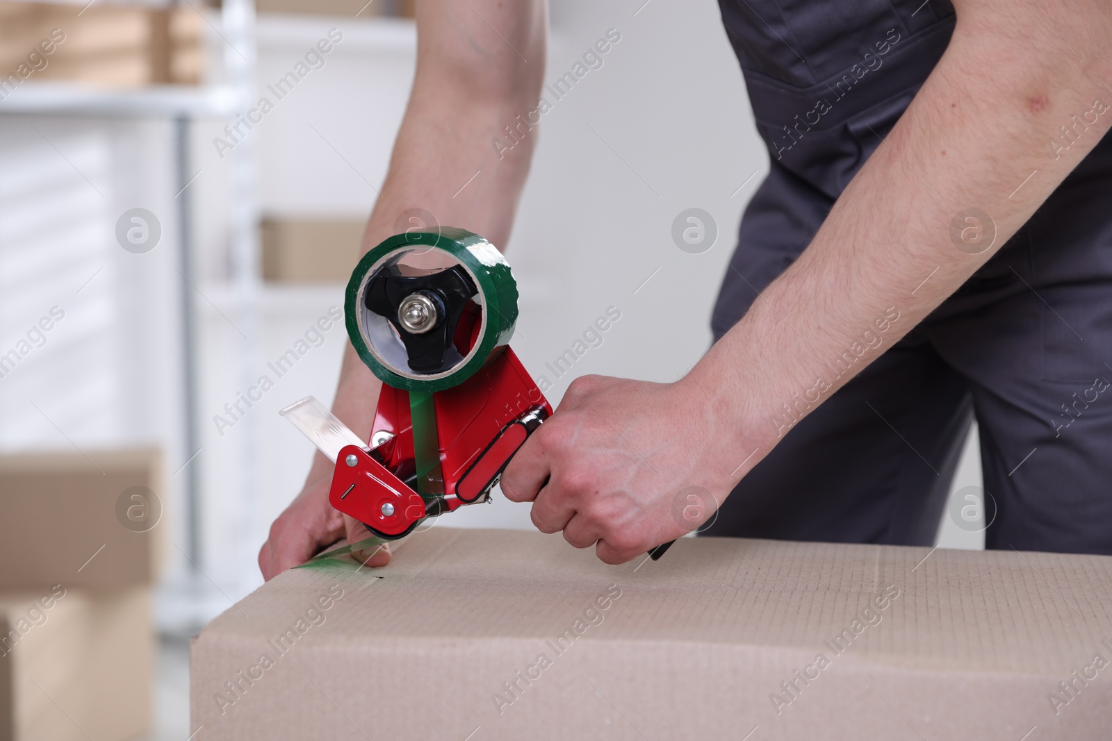 Photo of Man using dispenser with adhesive tape to seal cardboard box indoors, closeup