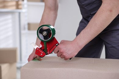 Photo of Man using dispenser with adhesive tape to seal cardboard box indoors, closeup