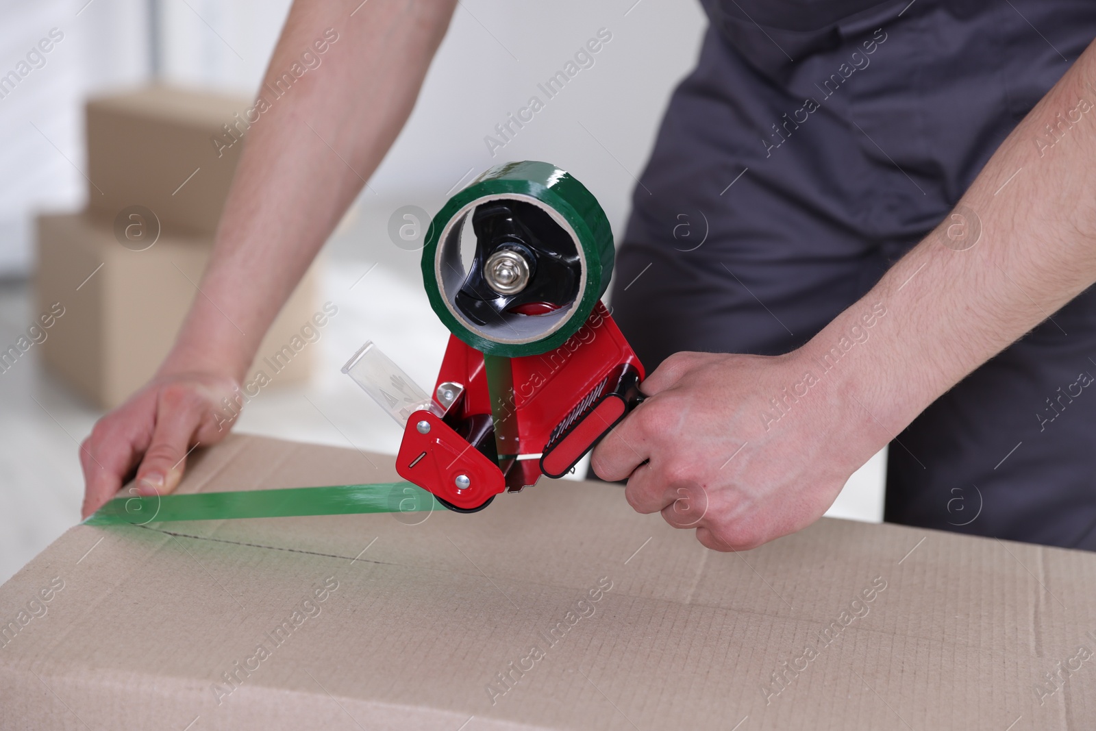 Photo of Man using dispenser with adhesive tape to seal cardboard box indoors, closeup