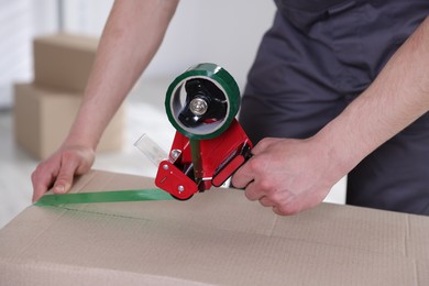 Photo of Man using dispenser with adhesive tape to seal cardboard box indoors, closeup