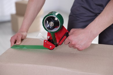 Photo of Man using dispenser with adhesive tape to seal cardboard box indoors, closeup