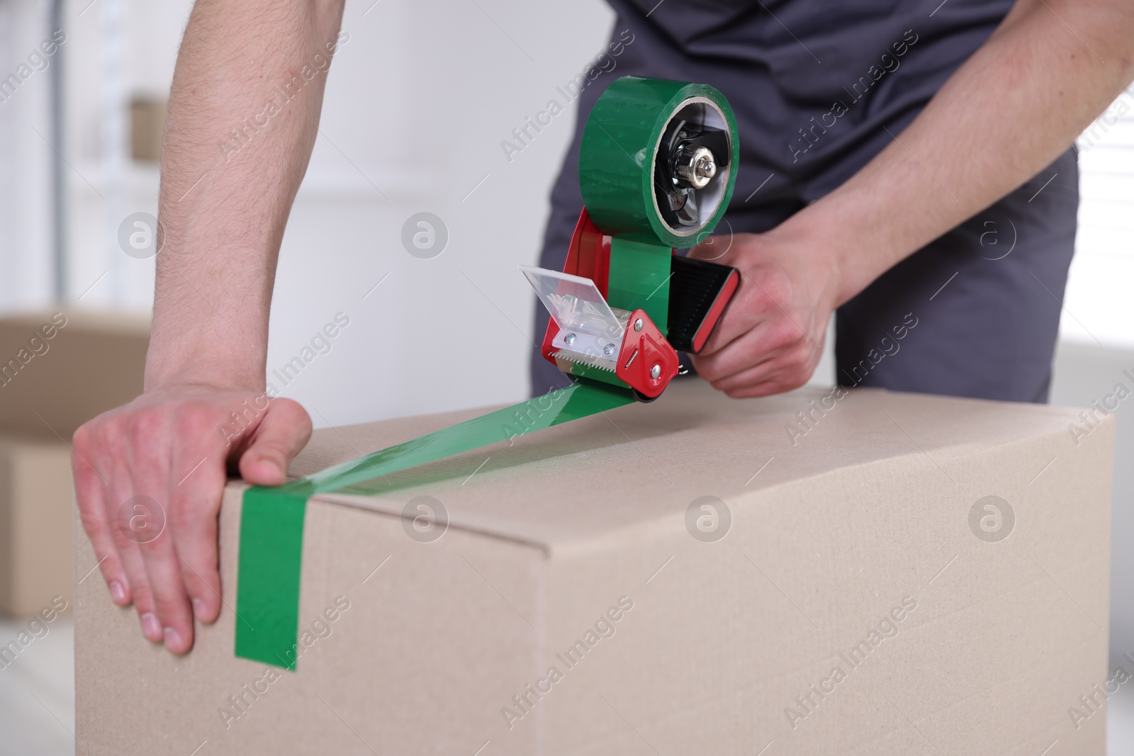 Photo of Man using dispenser with adhesive tape to seal cardboard box indoors, closeup