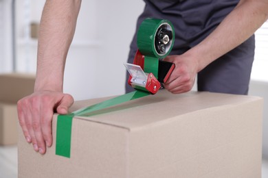 Photo of Man using dispenser with adhesive tape to seal cardboard box indoors, closeup