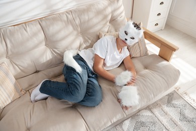 Photo of Quadrobics. Boy wearing cat mask, gloves and tail on sofa at home