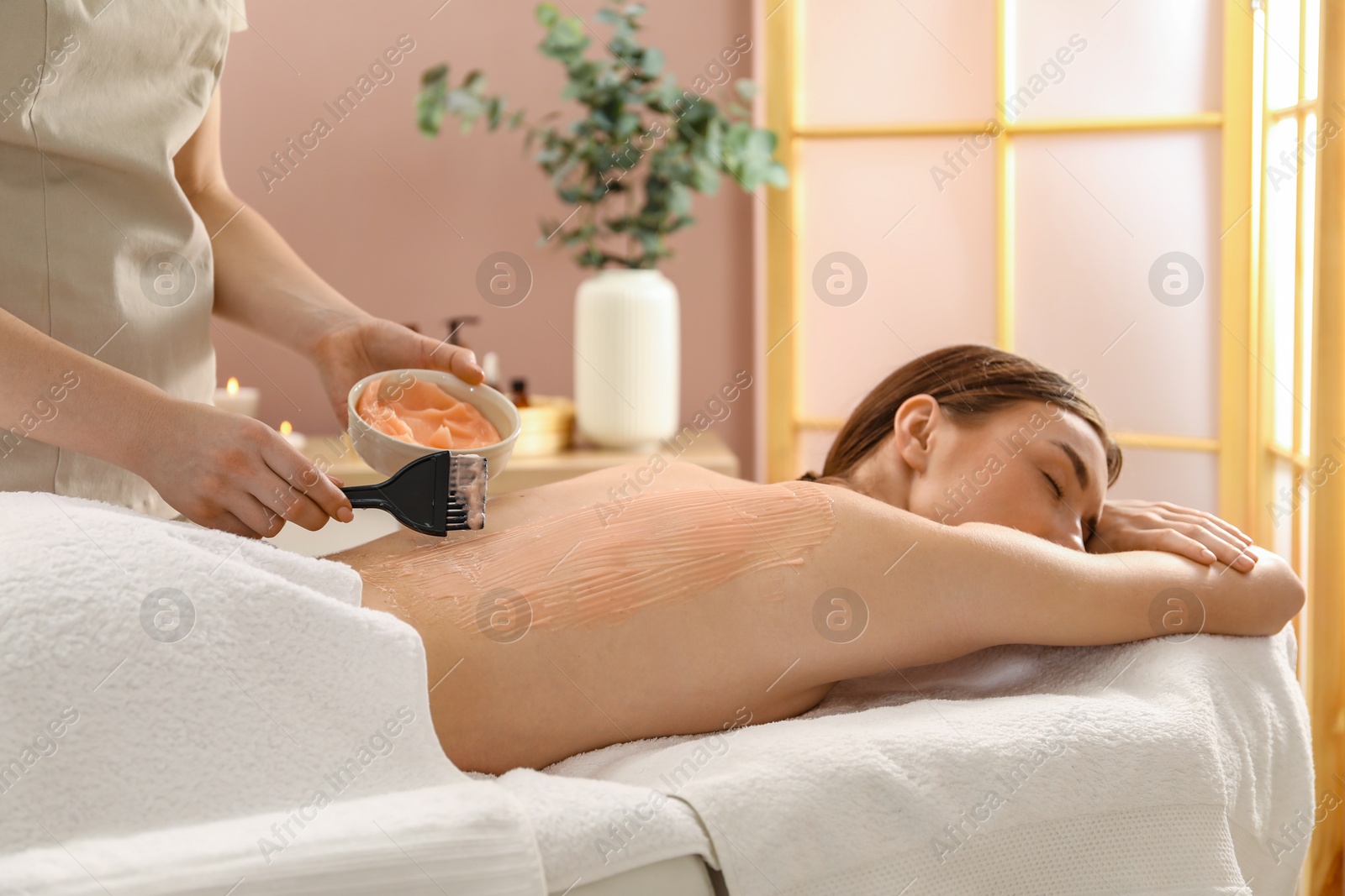 Photo of Esthetician applying cosmetic product for body wraps treatment onto woman's back in spa salon, closeup