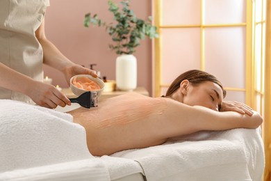 Photo of Esthetician applying cosmetic product for body wraps treatment onto woman's back in spa salon, closeup