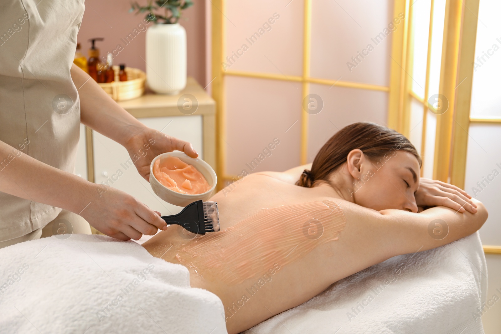 Photo of Esthetician applying cosmetic product for body wraps treatment onto woman's back in spa salon, closeup