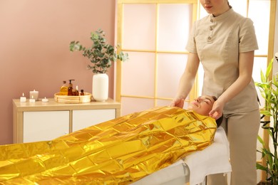 Photo of Spa body wraps. Esthetician covering woman with thermal foil blanket indoors, closeup. Space for text