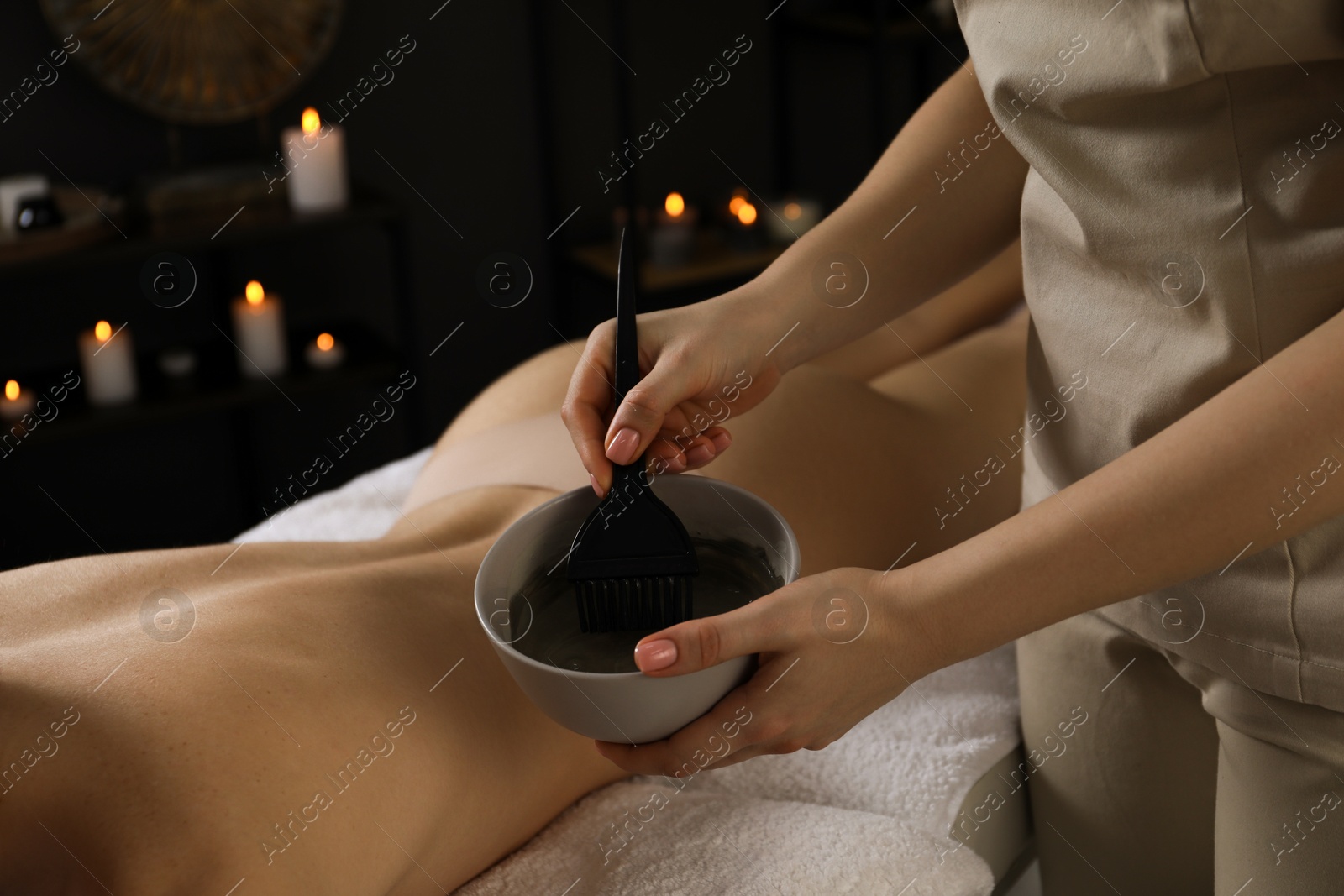 Photo of Esthetician applying cosmetic product for body wraps treatment onto woman's back in spa salon, closeup