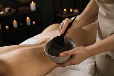 Photo of Esthetician applying cosmetic product for body wraps treatment onto woman's back in spa salon, closeup