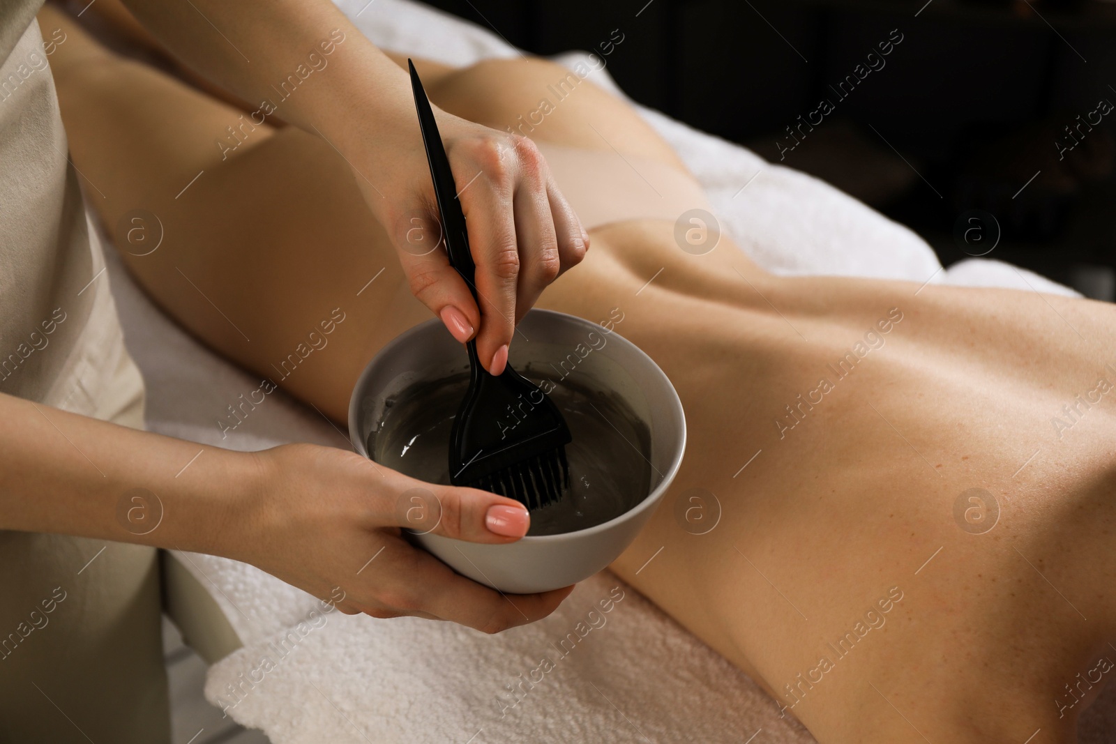 Photo of Esthetician applying cosmetic product for body wraps treatment onto woman's back in spa salon, closeup